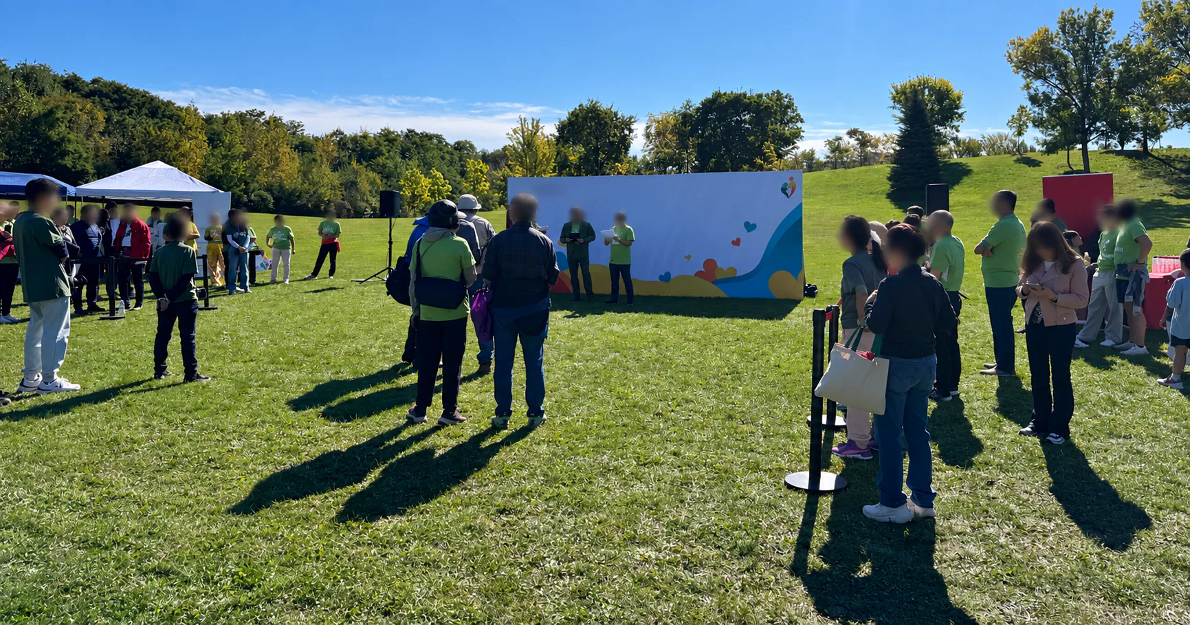 People gather beside a colourful event backdrop and speakers on a grassy park field.