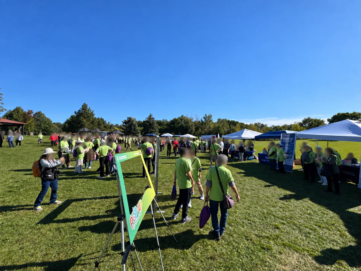 Participants in green shirts gather near tents and activity booths in a sunny park.